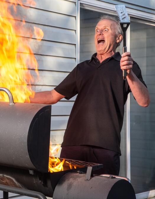 A man with wide eyes and open mouth holding a spatula looking a large flame on a grill